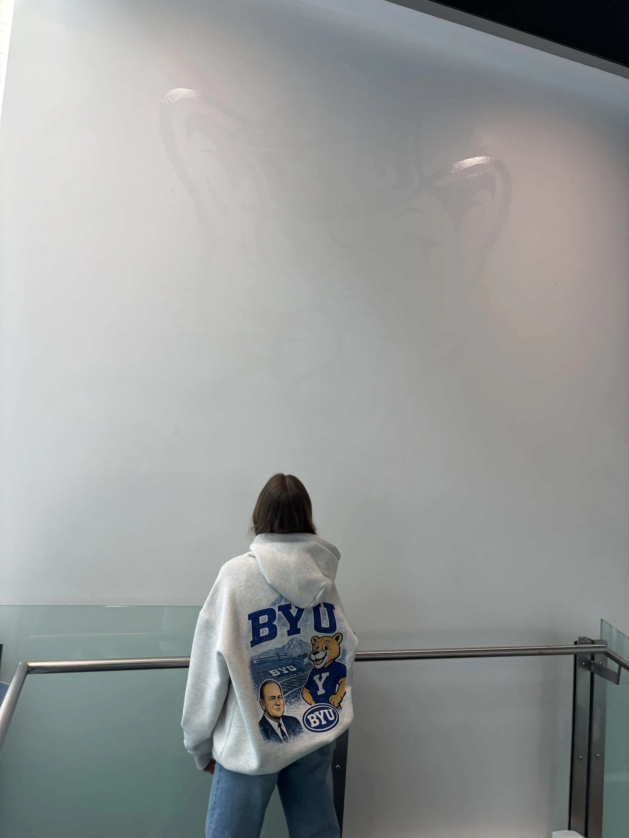 A person with shoulder-length hair wearing a gray hoodie with Yale University logos, standing in front of a light blue wall with a shadow of a coffee cup and saucer, looking away from the camera.