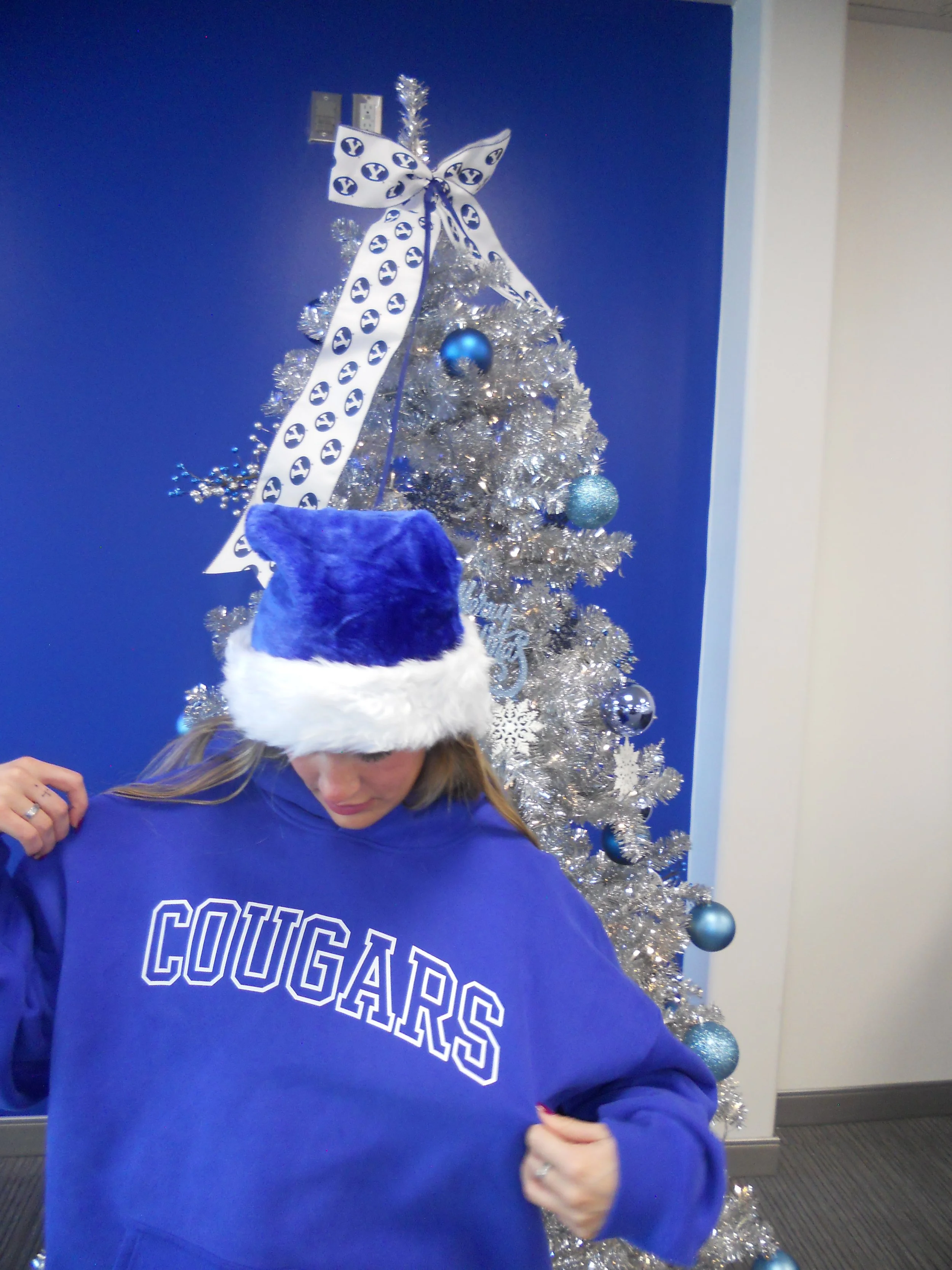 A woman wearing a blue sweatshirt with 'COUGARS' written on it and a Christmas hat with a blue and white design, standing in front of a silver Christmas tree decorated with blue and silver ornaments.