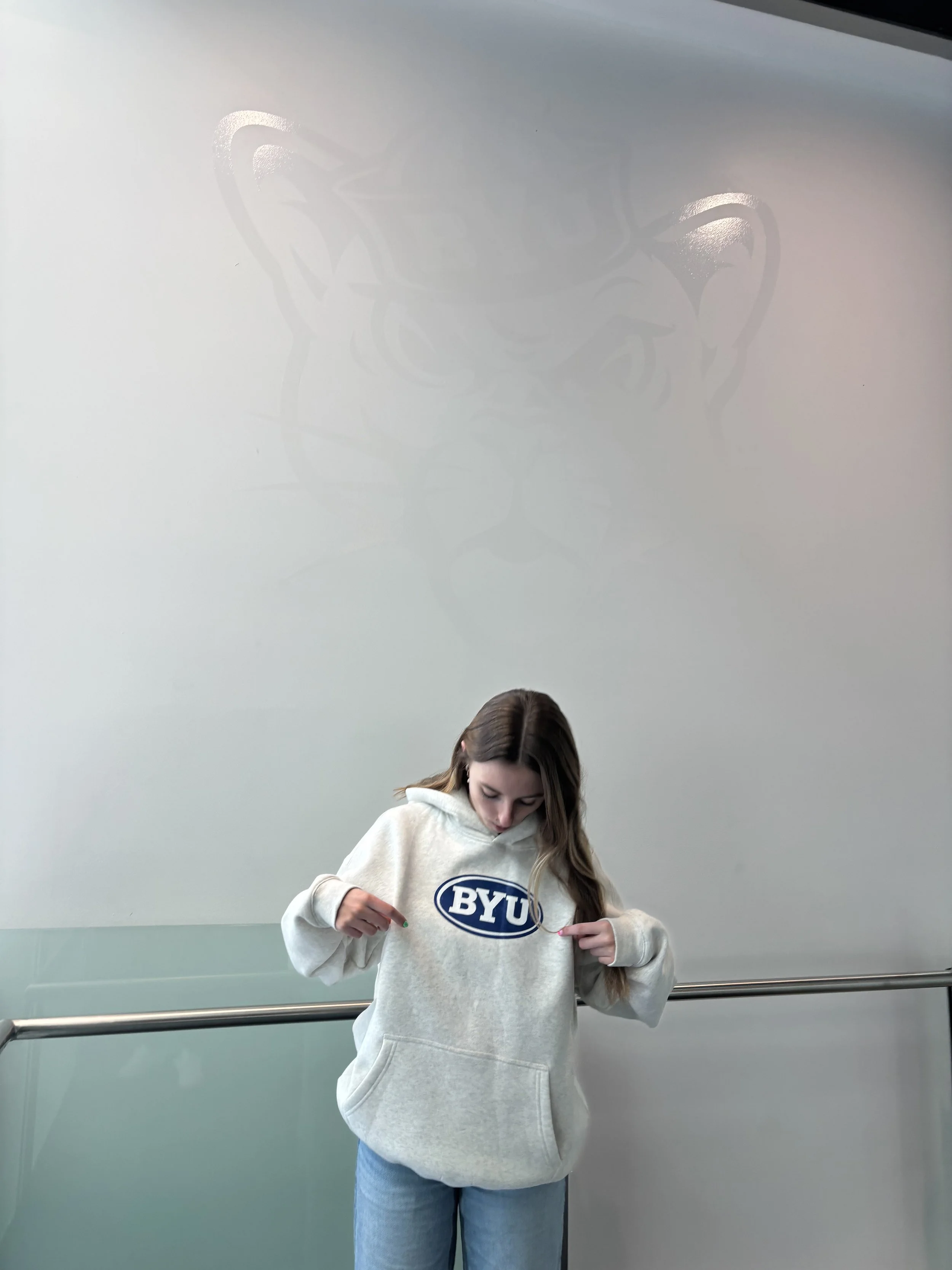 A young woman with long hair wearing a BYU sweatshirt standing in front of a plain wall with a reflection of a large decorative shadow or artwork overhead.