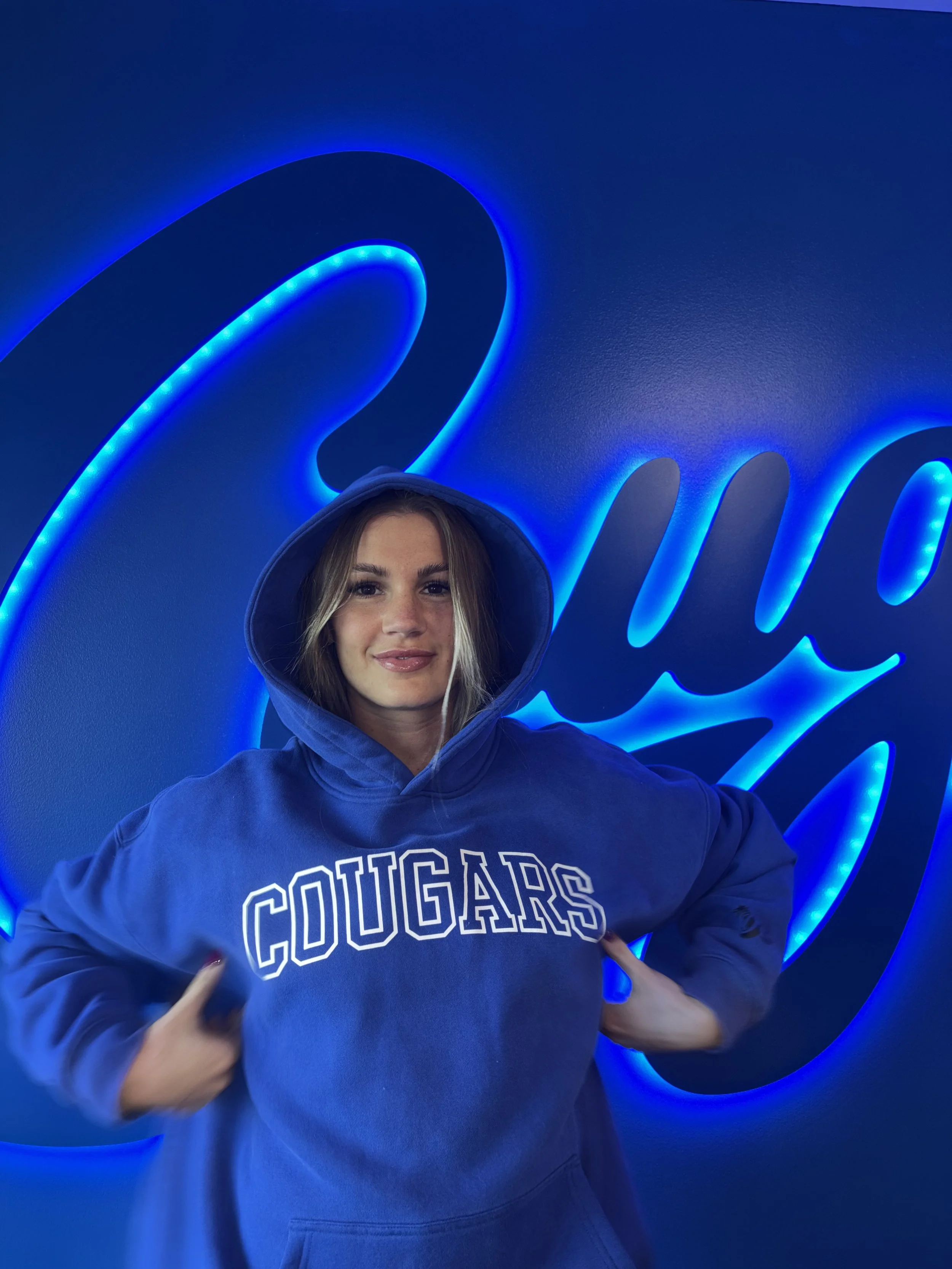 A young woman with long blonde hair wearing a blue hoodie with the word 'Cougars' on it, standing in front of a blue wall with illuminated blue neon cursive writing.