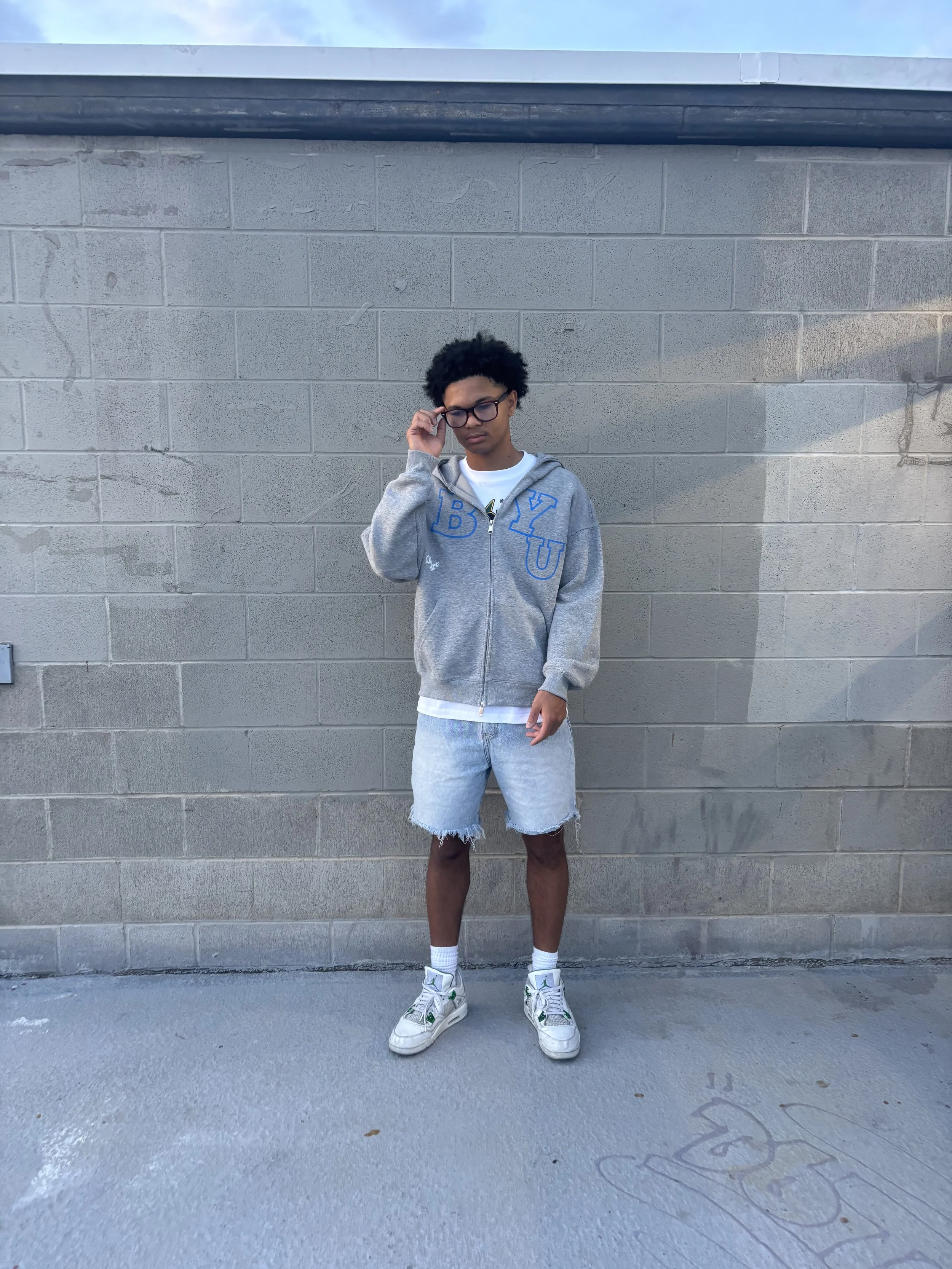 A young man with curly black hair and glasses stands against a gray concrete wall, wearing a gray hoodie with blue letters, shorts, and sneakers, adjusting his glasses.