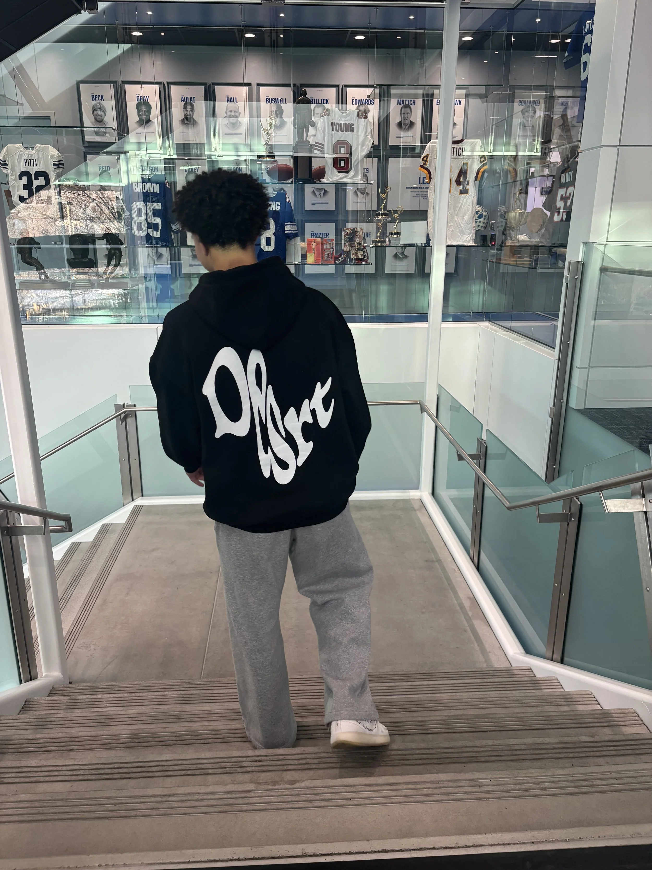 A person with curly hair wearing a black hoodie with white text on the back, gray sweatpants, and white sneakers, kneeling on a stairway inside a building with glass walls and display cases containing sports jerseys, trophies, and framed photos.