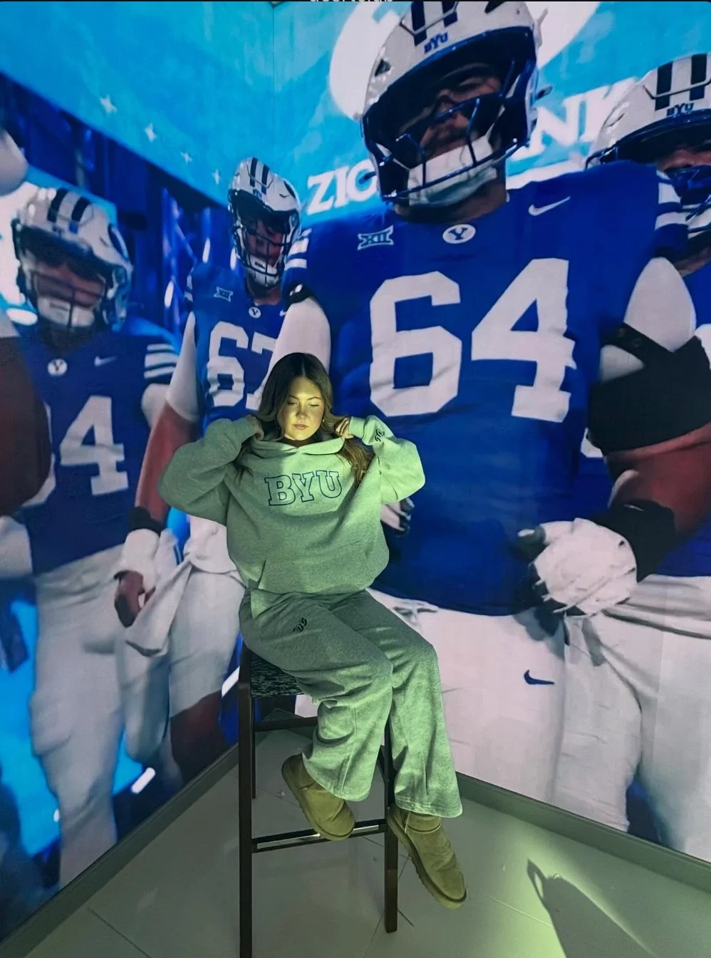 A woman sitting on a stool with her eyes closed, wearing a gray BYU sweatshirt and pants, in front of a large poster of football players in blue BYU uniforms.