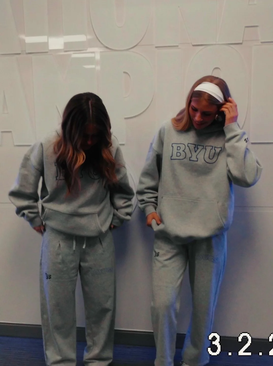 Two women in gray BYU sweatshirts and sweatpants standing with their heads bowed, facing downward, in front of a wall with large white letters. One woman has long wavy hair, the other has straight hair with a white headband. The photo has a timestamp