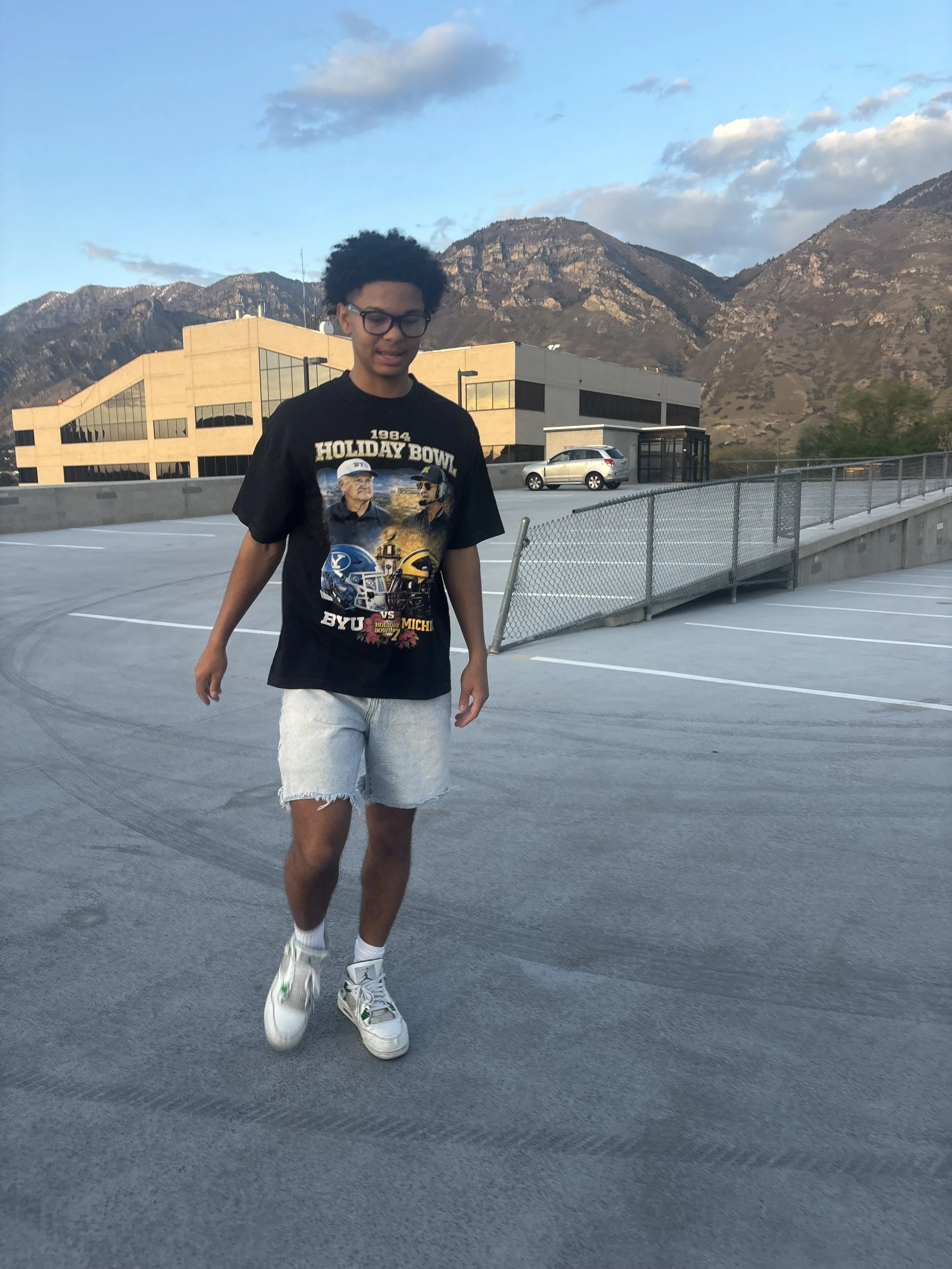 A young man with curly hair, glasses, and light-colored shorts walking on a parking garage rooftop, with mountains and a blue sky with clouds in the background.