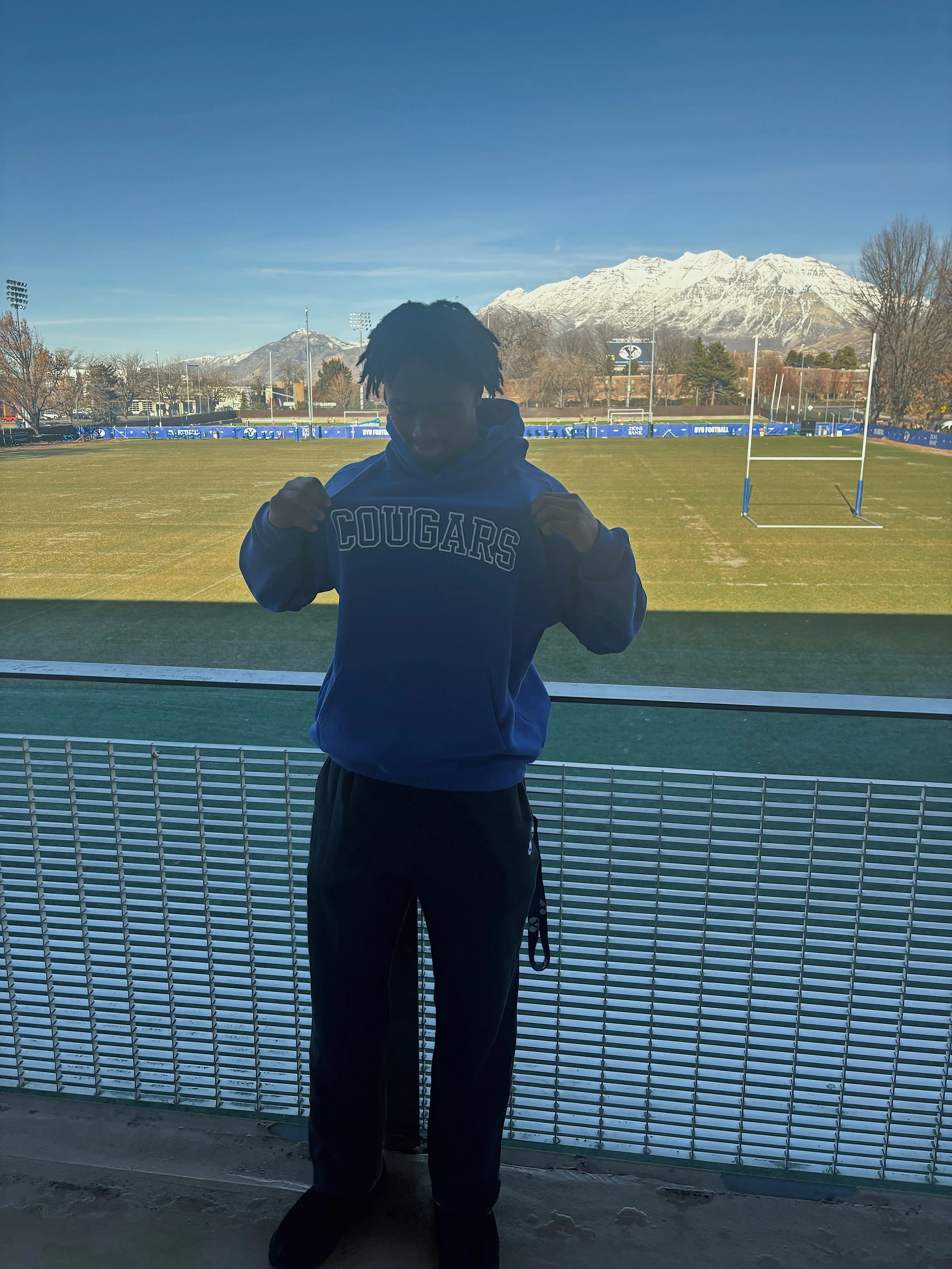 Person standing behind a glass window at a sports field, wearing a blue hoodie with 'COUGARS' written on it, with snow-capped mountains in the background.