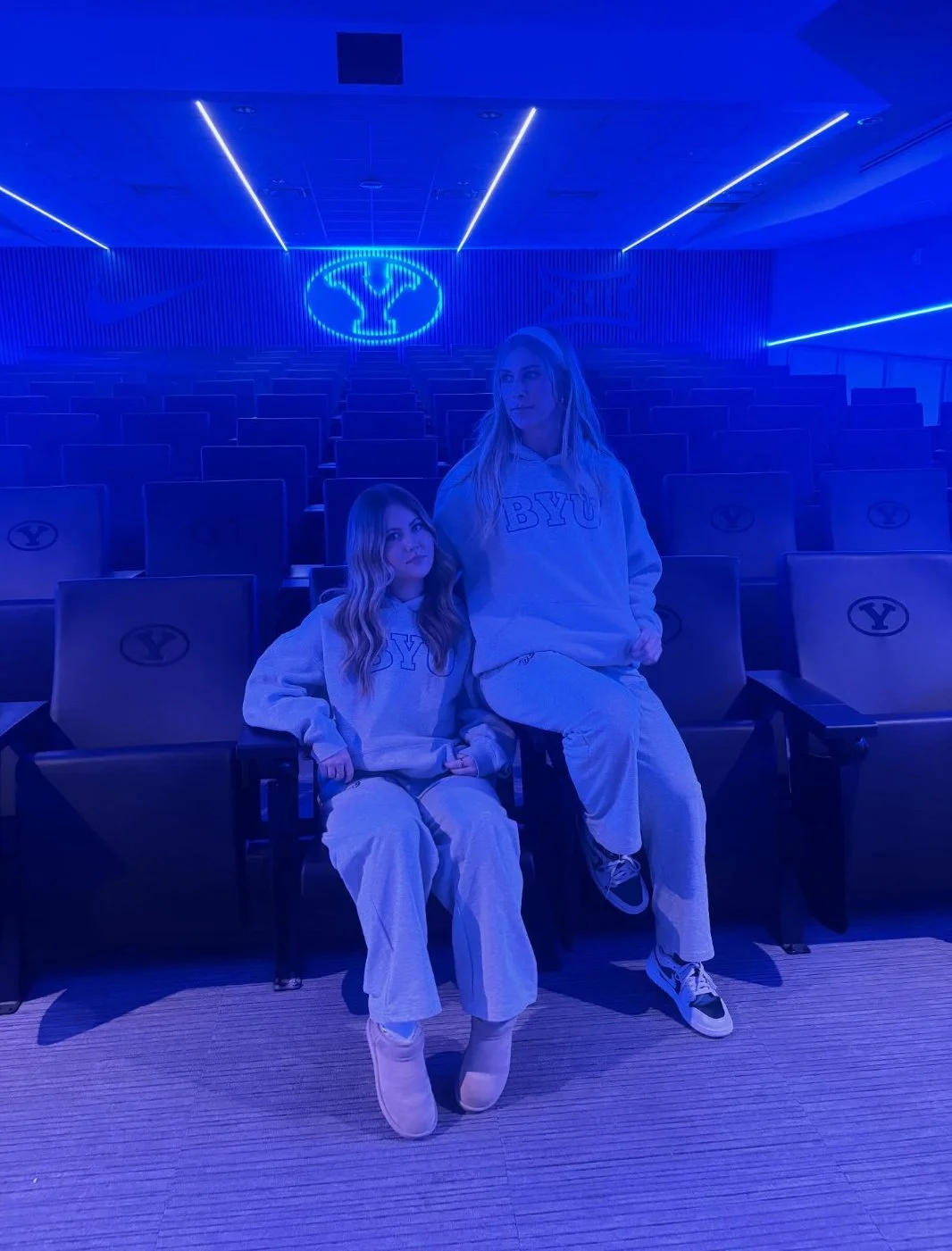 Two women wearing BYU sweatshirts sitting and standing in a dimly lit, blue-tinted stadium or auditorium with empty seats and a glowing BYU logo on the wall behind them.