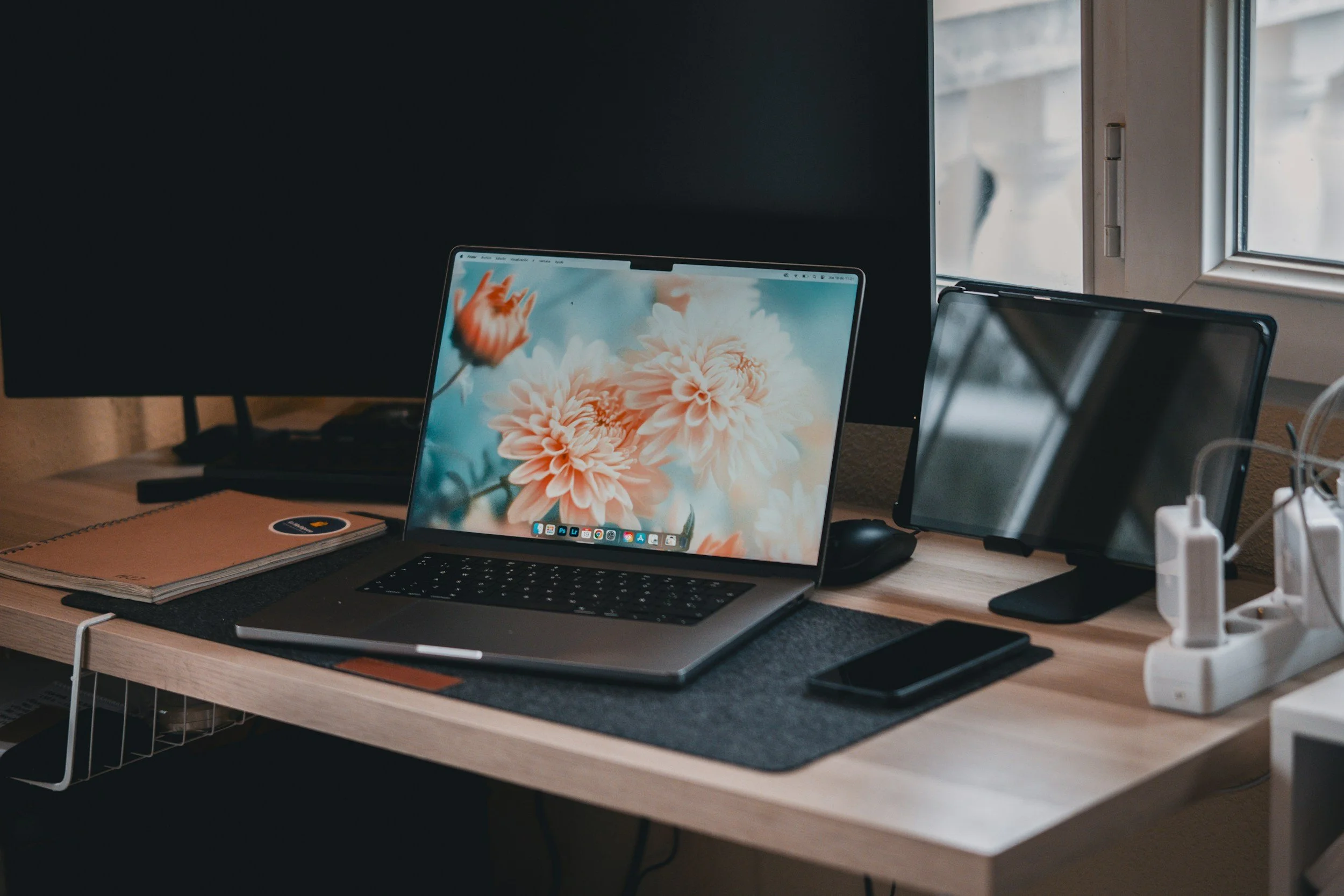 Computer workspace with a laptop, monitor, tablet, smartphone, notebooks, and charging cables on a wooden desk.