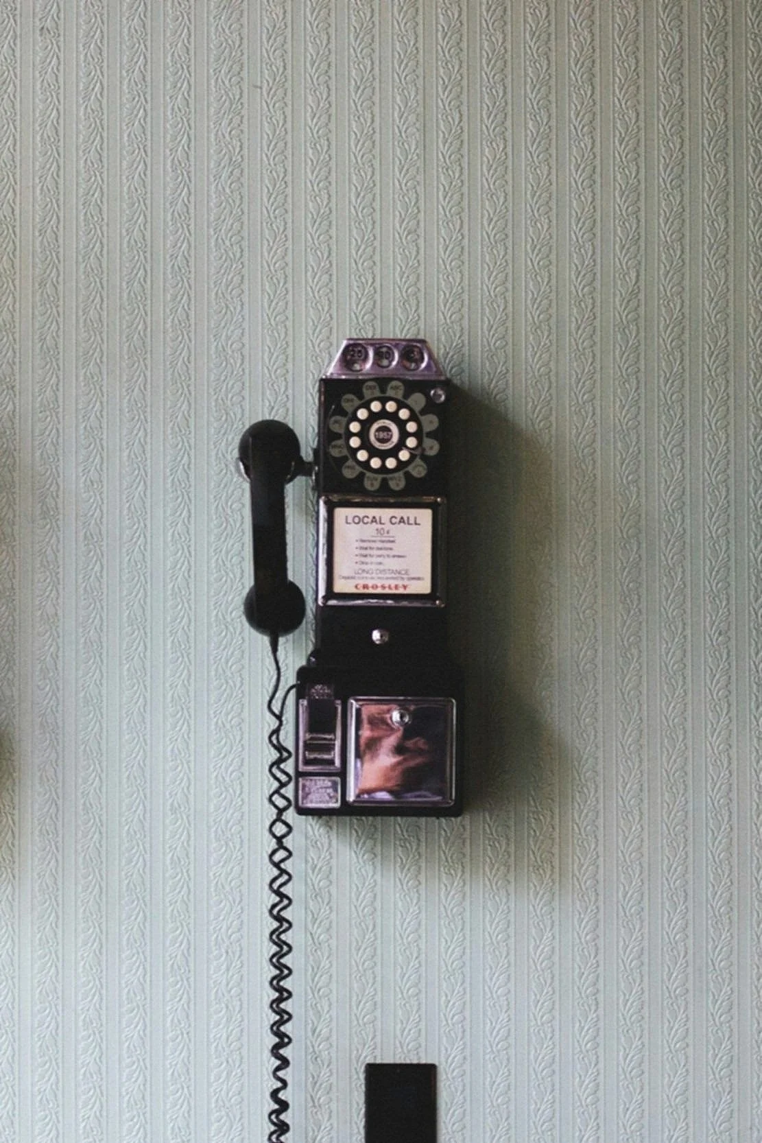 Vintage black rotary wall phone mounted on a green patterned wallpaper wall.