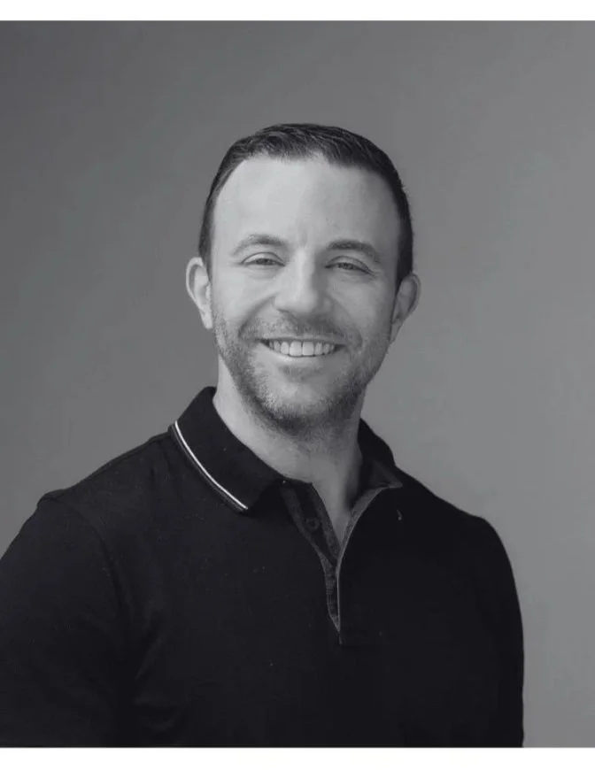 Black and white photo of a man smiling slightly with short hair, wearing a dark shirt, against a plain background.