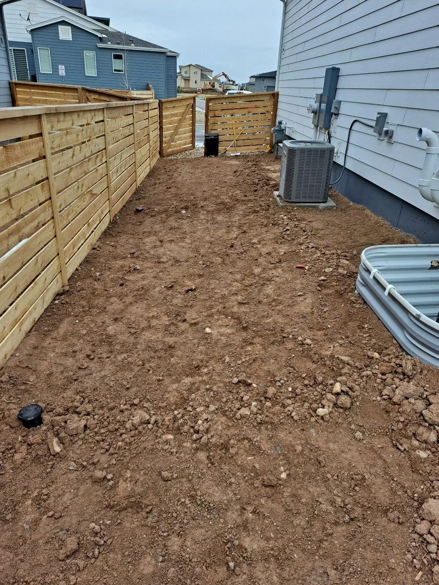 A backyard with dirt ground, wooden fence on the left, white house on the right, and construction equipment in the background.