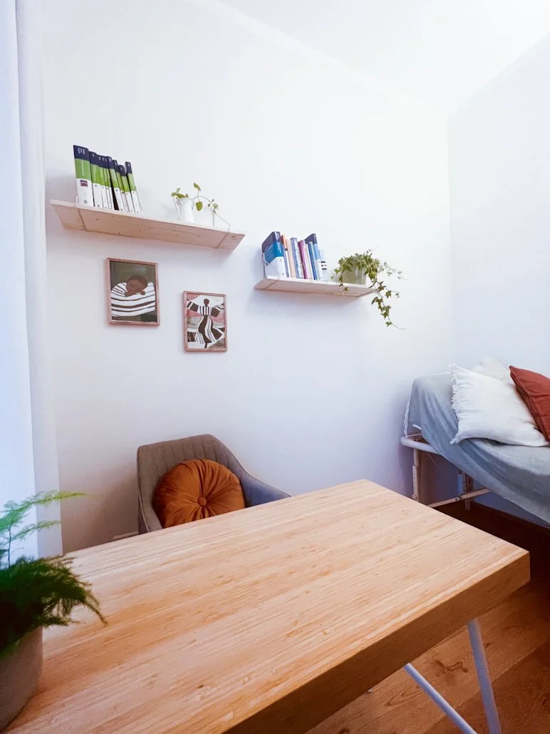 Ein gemütliches Zimmer mit einem Holzschreibtisch, einem grauen Sessel mit braunem Kissen, einem Bett mit weißen und roten Kissen, und an der Wand hängen zwei Bilder. Es gibt auch Wandregale mit Büchern und Pflanzen.