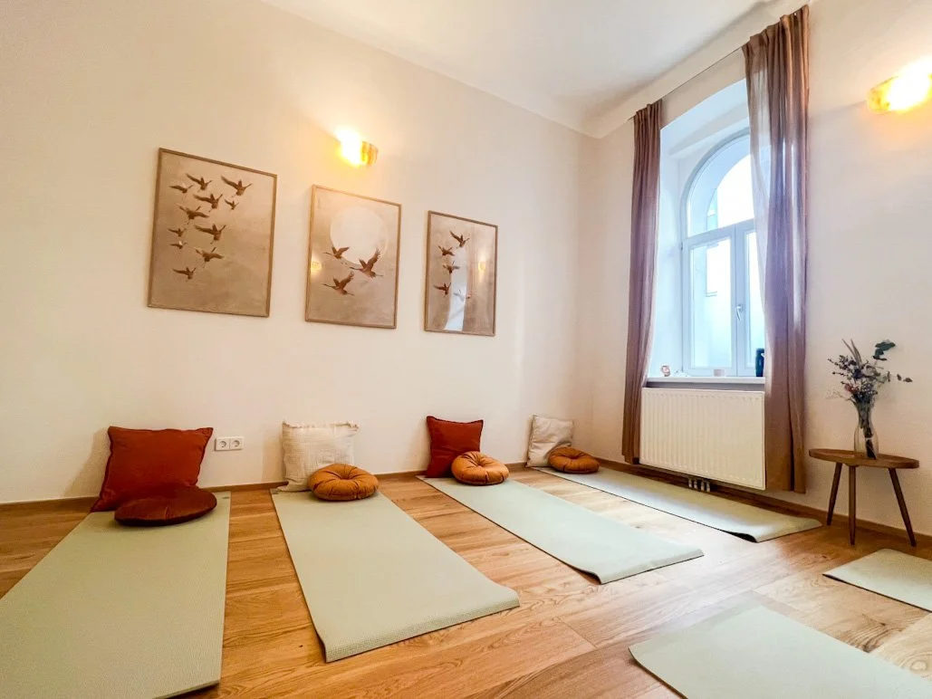 Yogaraum mit fünf Yogamatten und langen Kissen, Wanddekoration mit Vogelbildern, Fenster mit Vorhängen, Tisch mit Blumen, warmes Licht.