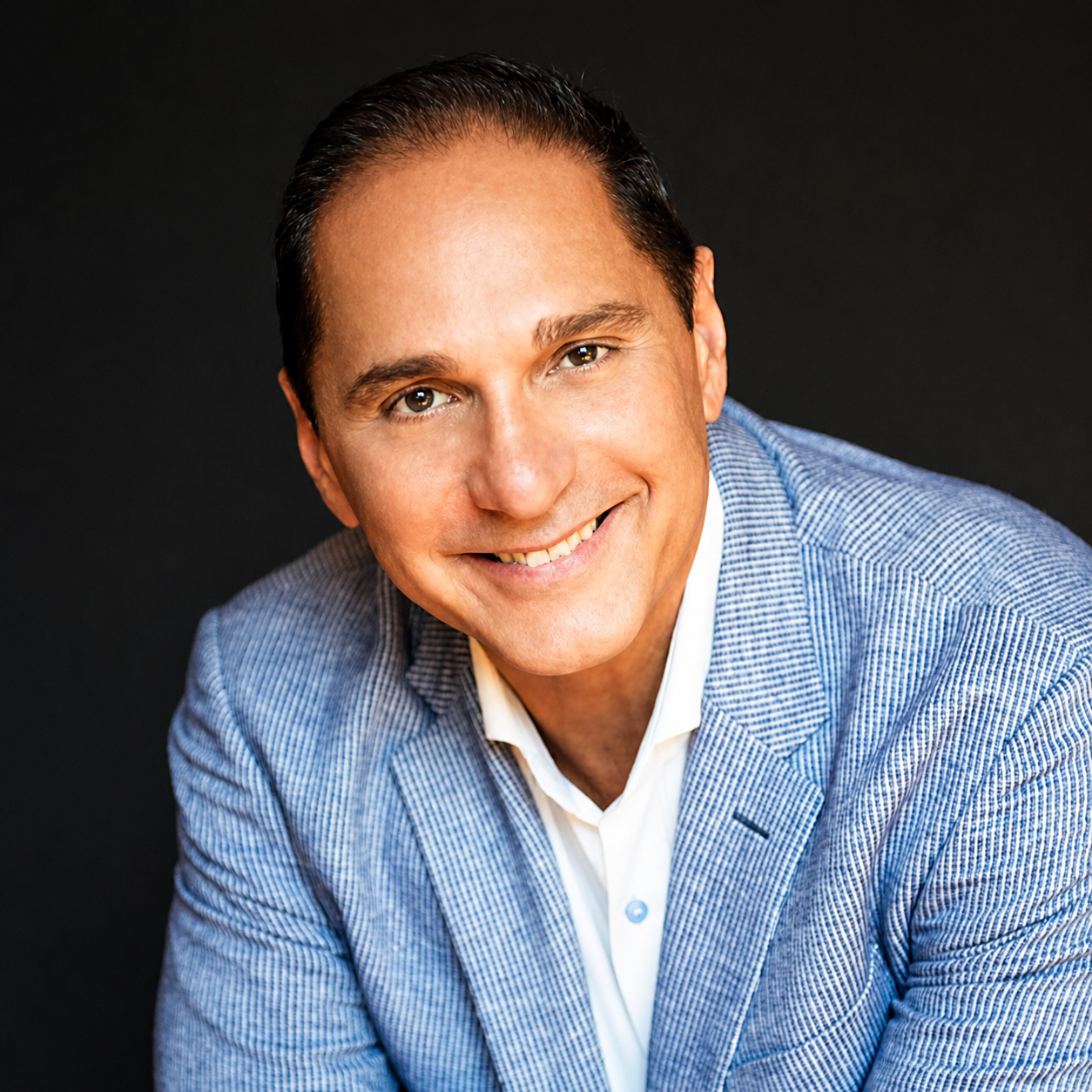 Portrait of a smiling man in a blue blazer and white shirt against a dark background.