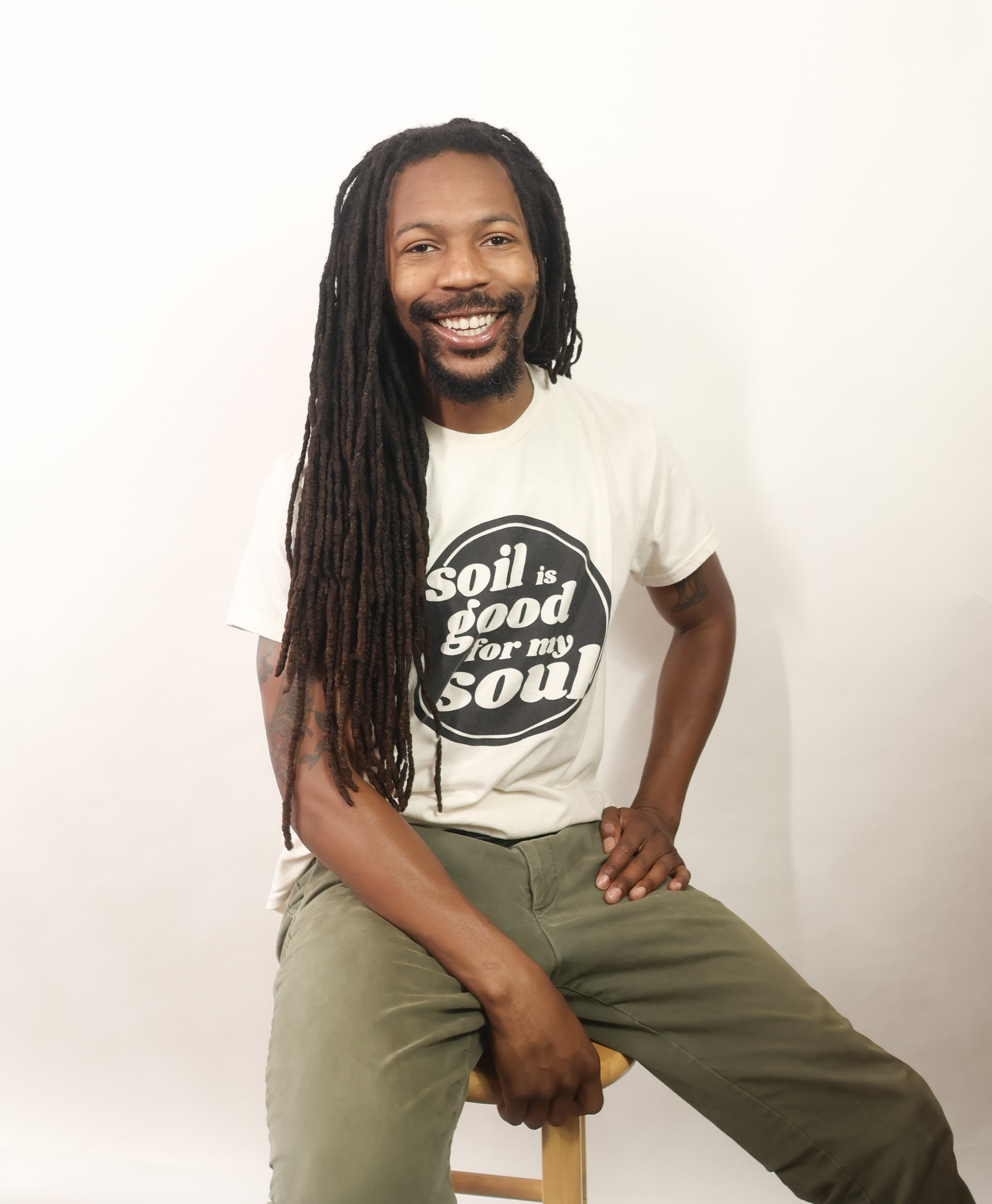 A smiling man with long dreadlocks and a beard, wearing a white T-shirt with a black circle design and text, sitting on a stool against a plain white background.
