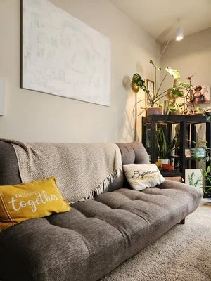 Living room with a gray couch, decorative pillows, a large wall art, and a plant shelf in the corner.