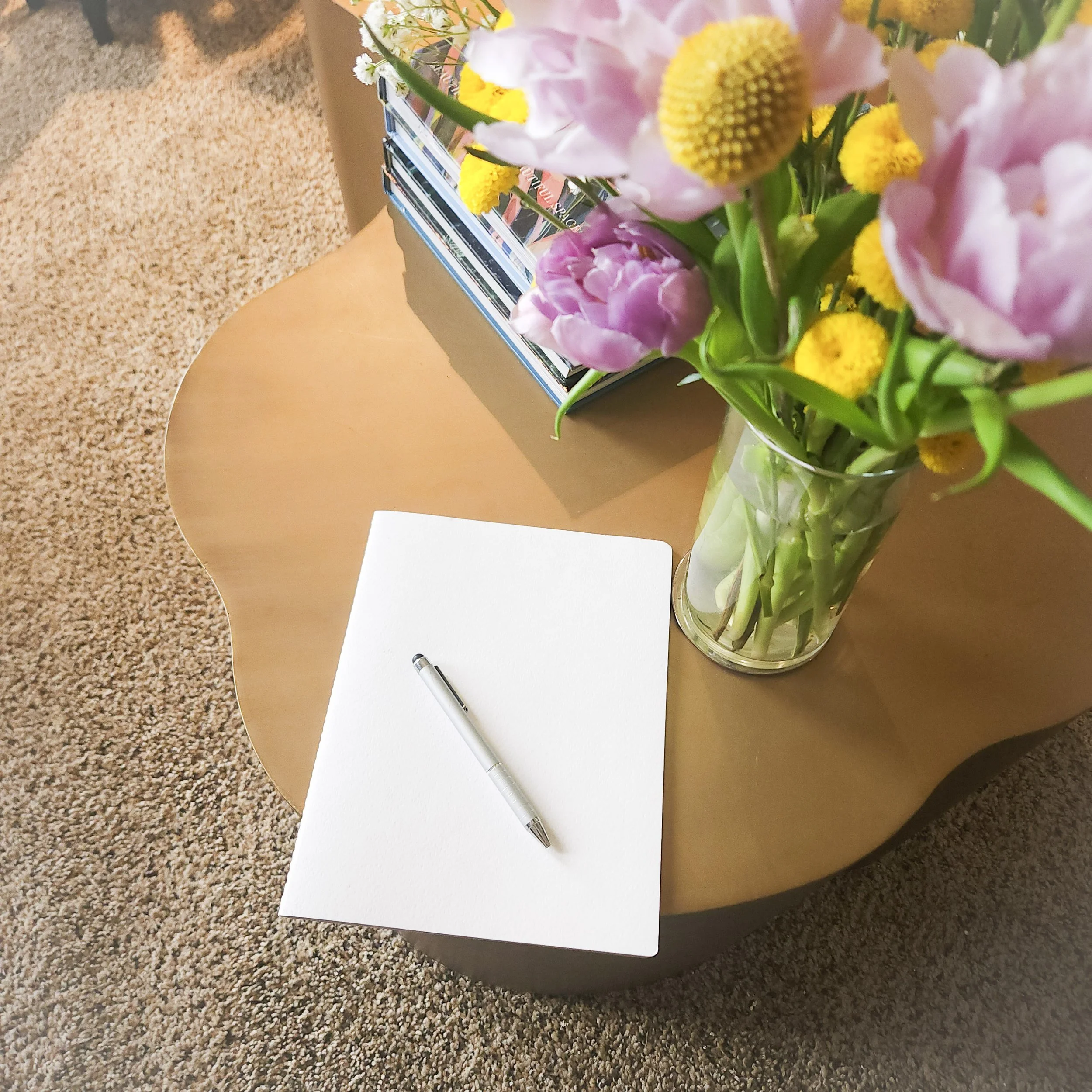 A bouquet of pink, white, and yellow flowers in a clear glass vase on a table, with a white piece of paper, a black pen, and a few magazines in the background.