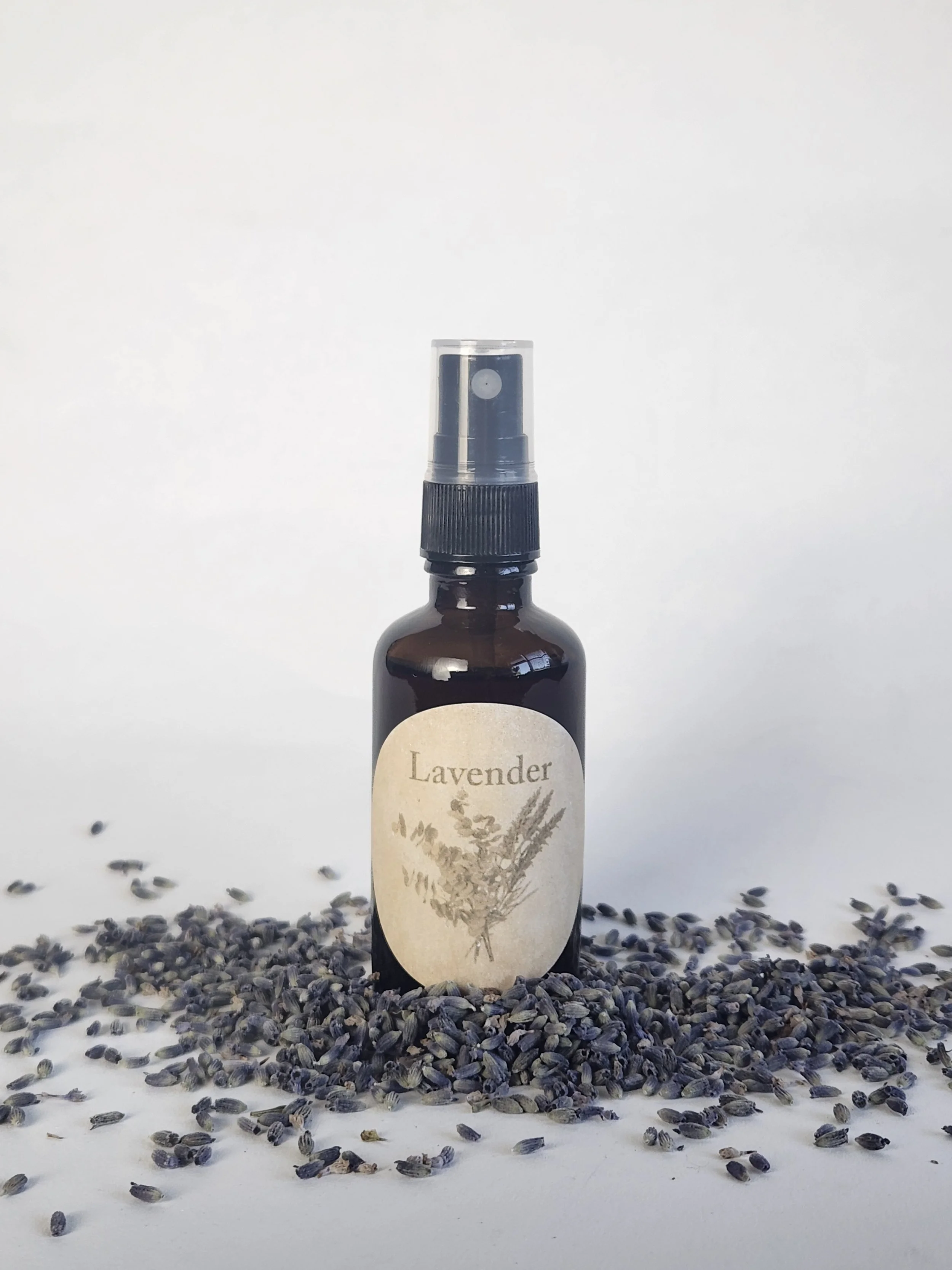 A brown glass spray bottle with a lavender label featuring an illustration of lavender flowers, standing among scattered lavender buds on a white background.
