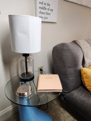 Glass side table holding a lamp with white shade, a set of silver coasters, and a pink notebook, next to a gray sofa with a yellow pillow in a living room.