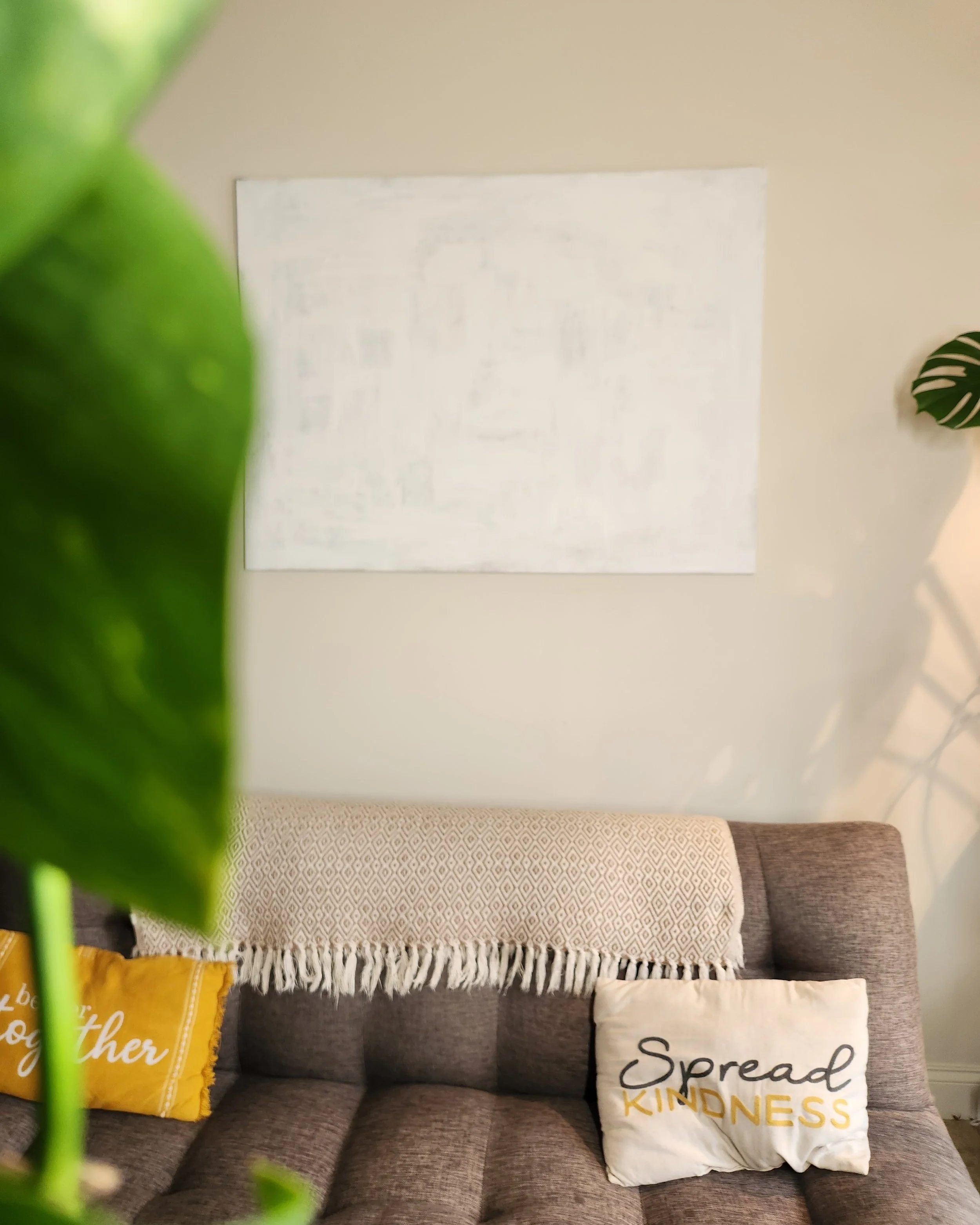 Living room with a grey sofa, white decorative pillow with black text reading 'Spread Kindness' in gold, yellow and white pillows, white wall with a large framed artwork, green plants partially visible in the foreground.