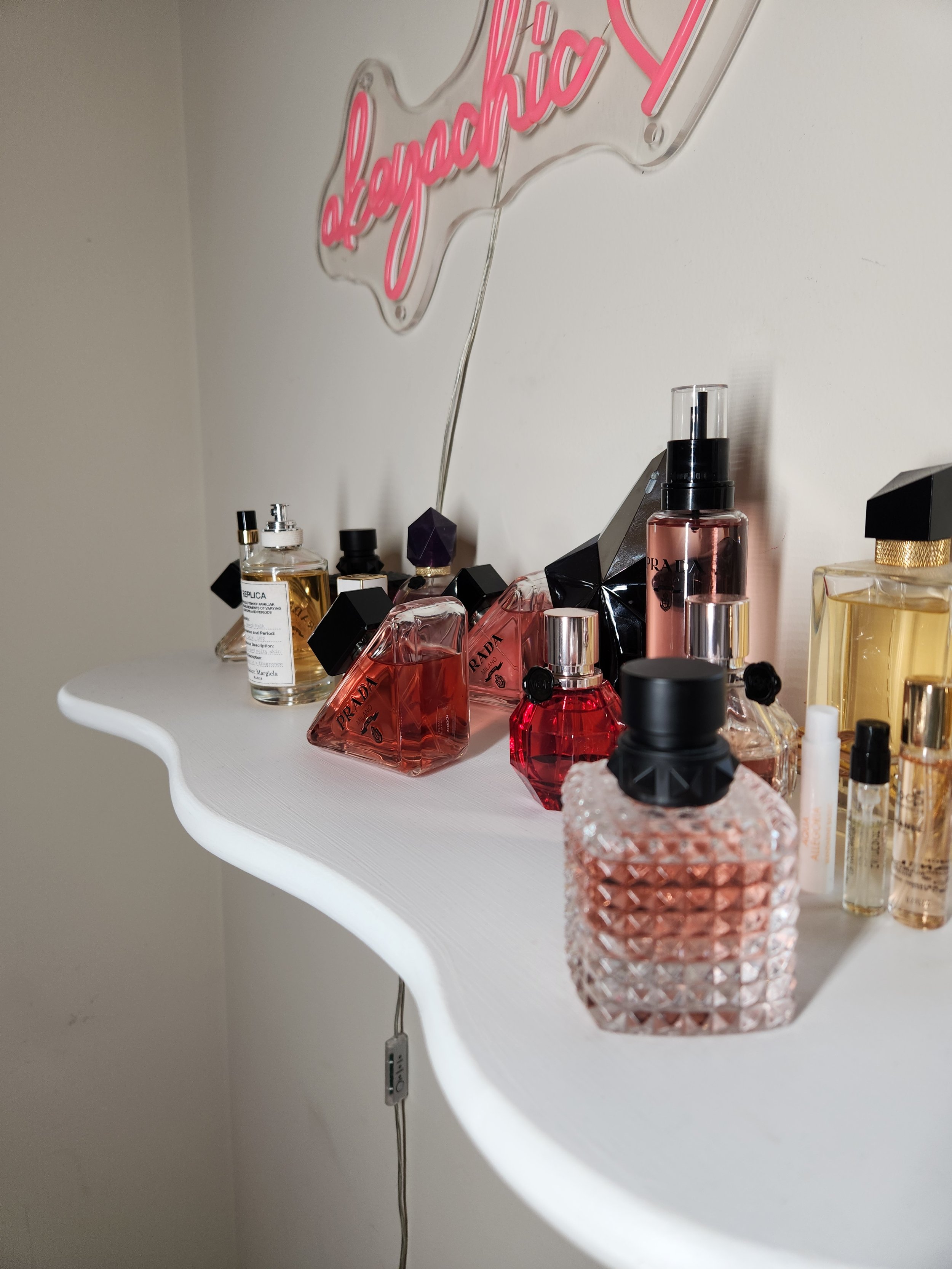 A white shelf holding various perfume bottles and a neon sign mounted on the wall behind the shelf.