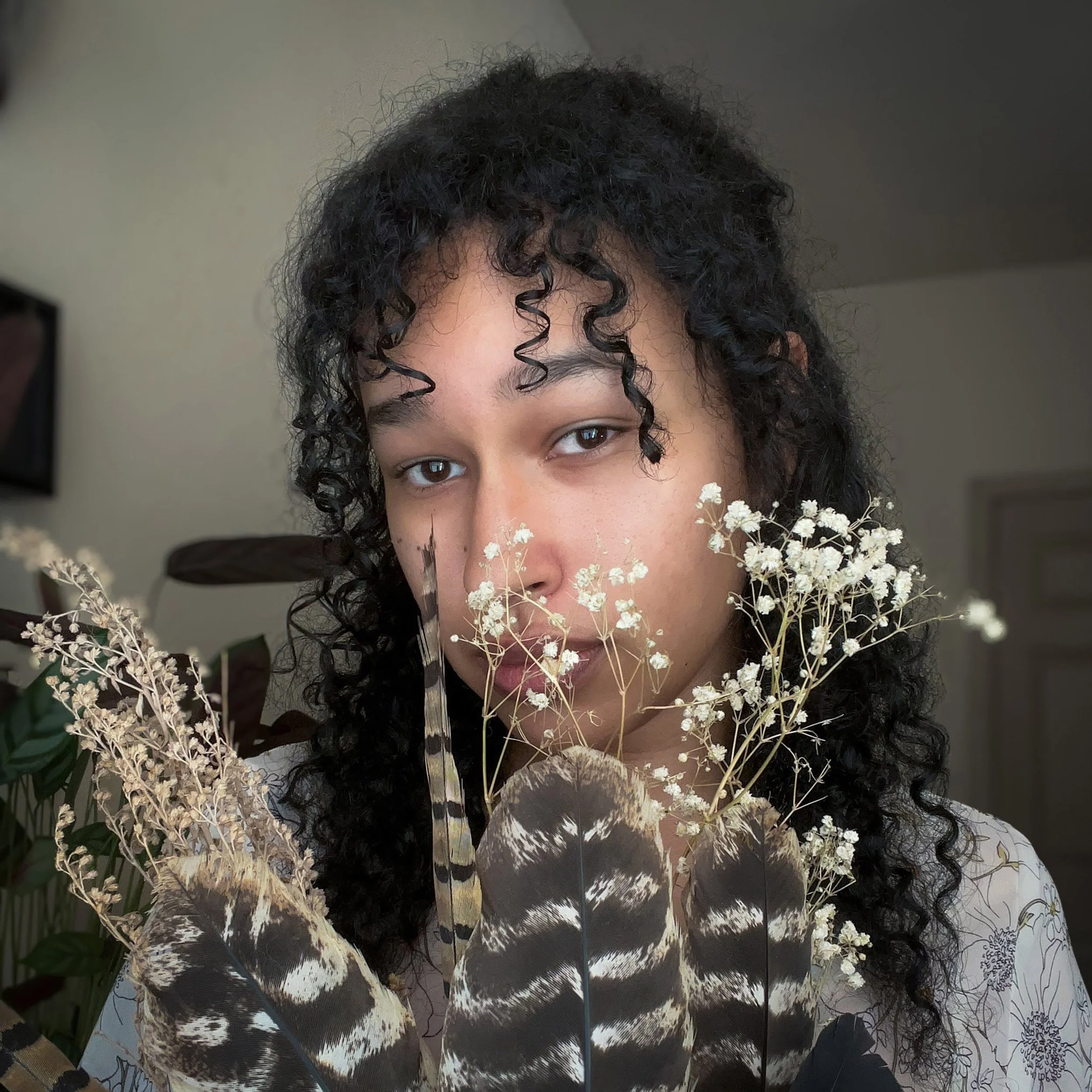 A person with curly dark hair holding feathers and white baby's breath flowers, looking at the camera indoors.