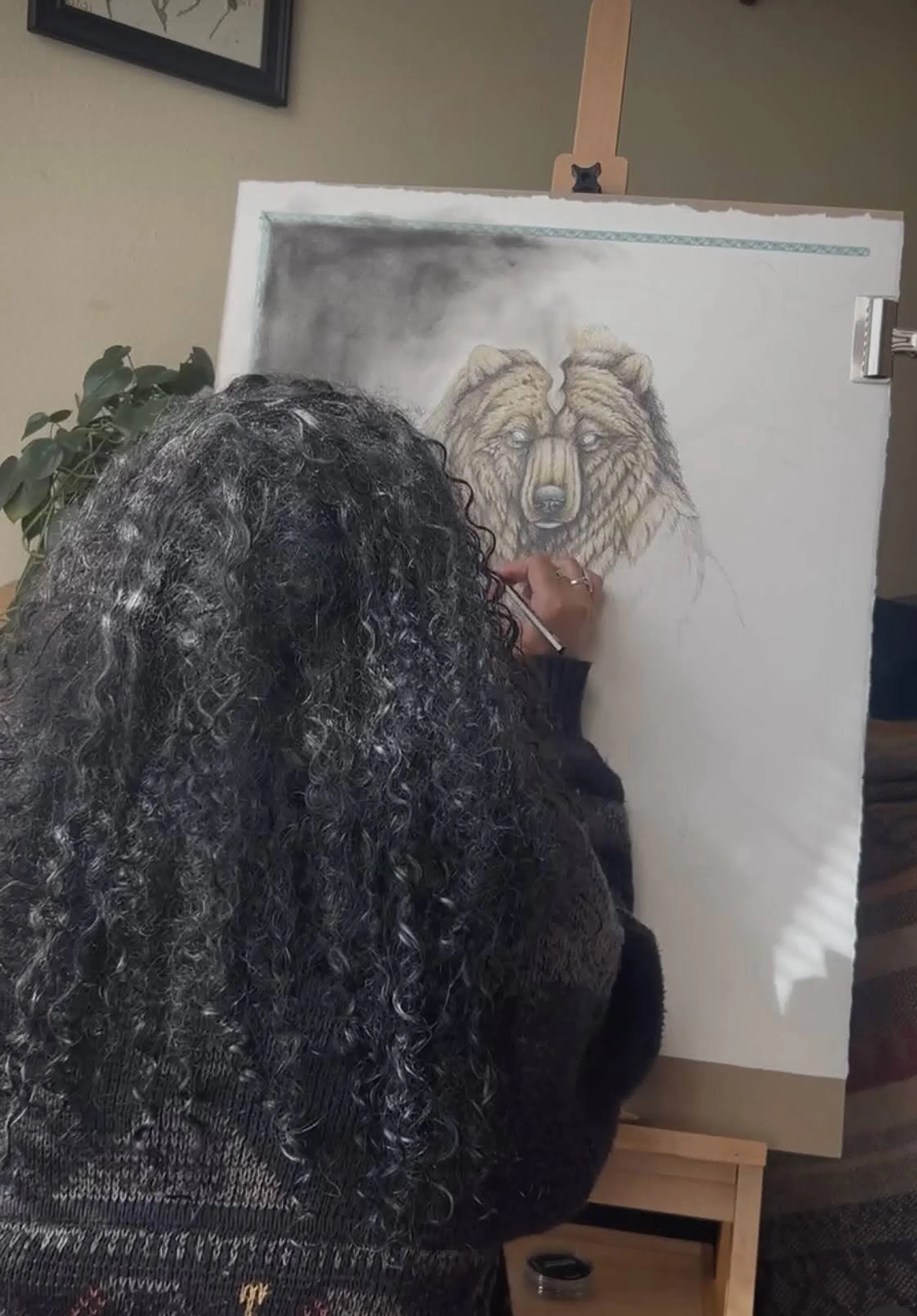 A person with curly hair sketching a realistic drawing of a bear on a canvas. The bear's face is detailed with a focus on its eyes and fur. There is a leafy plant to the left and a partially visible picture frame on the wall behind.