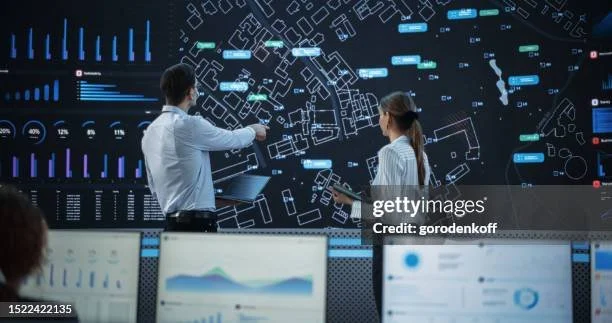 Two individuals in a control room analyzing digital maps and data visualizations on large screens, with multiple monitors displaying charts and graphs.