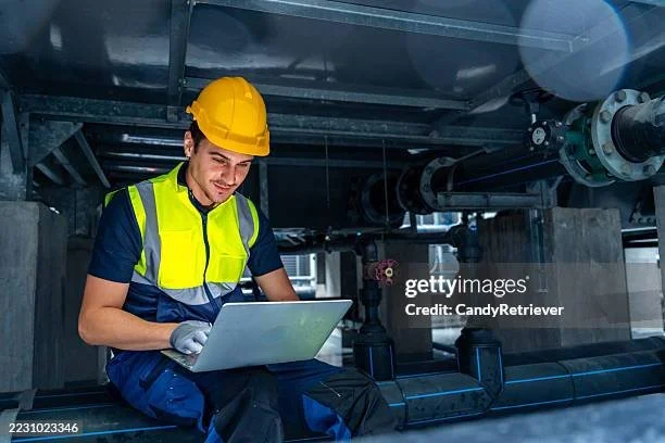 A male industrial worker wearing a yellow safety helmet and reflective vest, sitting under machinery, using a laptop.