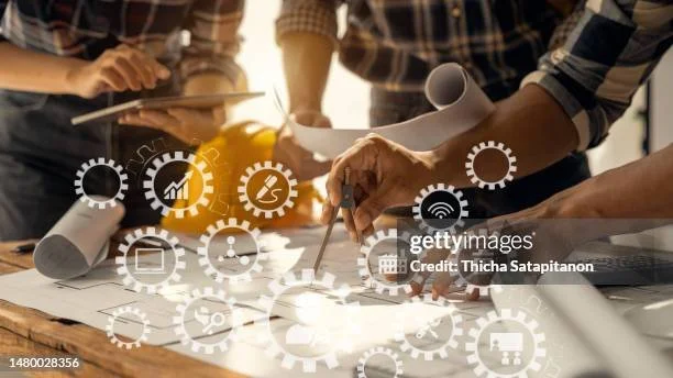 Group of people working together on a project, discussing plans with rolled-up blueprints on a table, with digital icons overlayed.