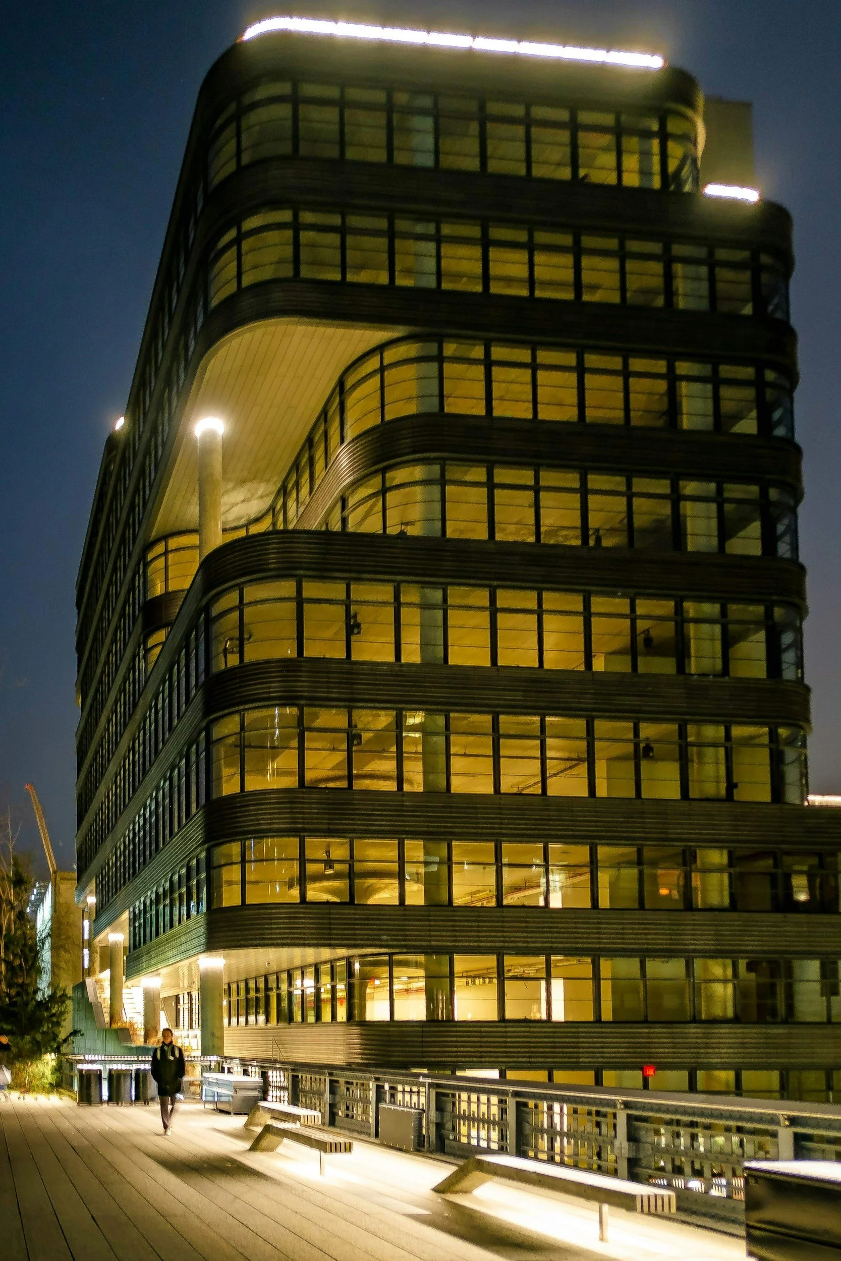 Nighttime view of a modern, multi-story building with curved design, large glass windows, and illuminated interior, with a person walking on a bridge-like pathway in the foreground.