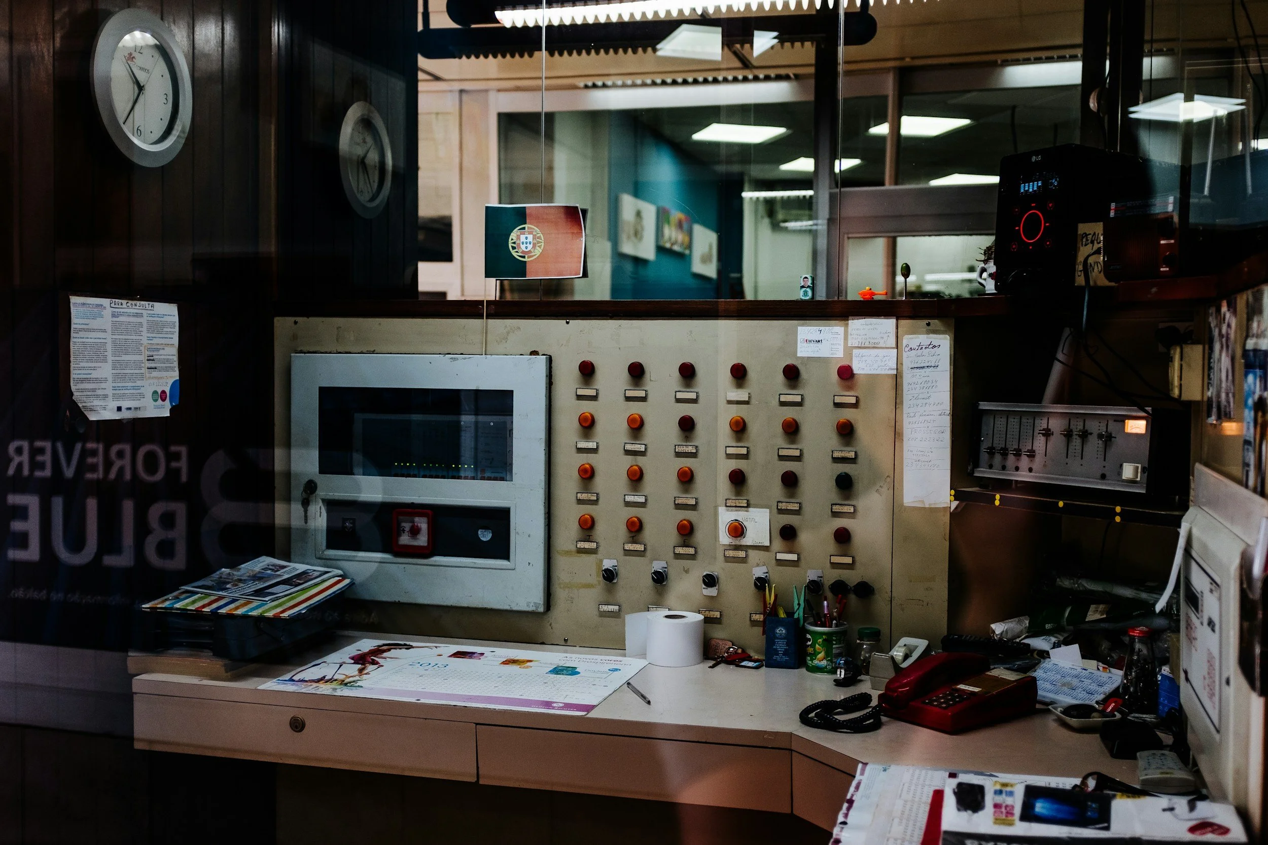 An indoor office control room with an array of switches, buttons, a monitor, and various office supplies on the desk. Two wall clocks and a small flag of Portugal are visible. There is a large window separating this room from another office space with ceiling lights.