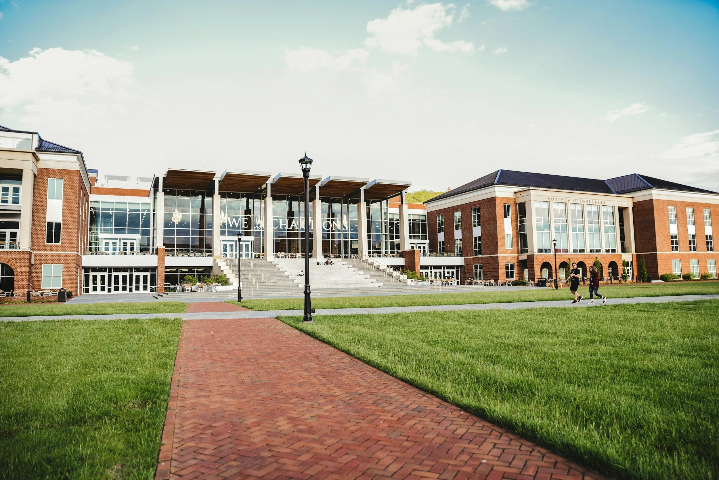 A college campus with a large modern building that has glass walls and brick exterior, with students walking on the green grass and brick pathway.