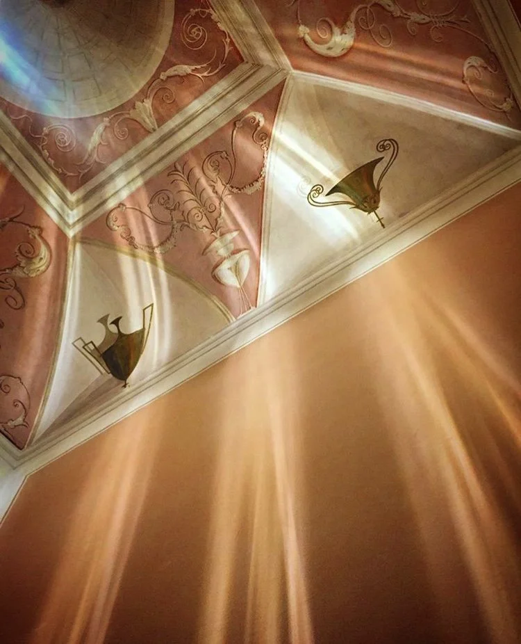 Ceiling with ornate pink and cream designs, gold accents, decorative wallpaper, and wall-mounted lanterns, with sunlight streaming in.