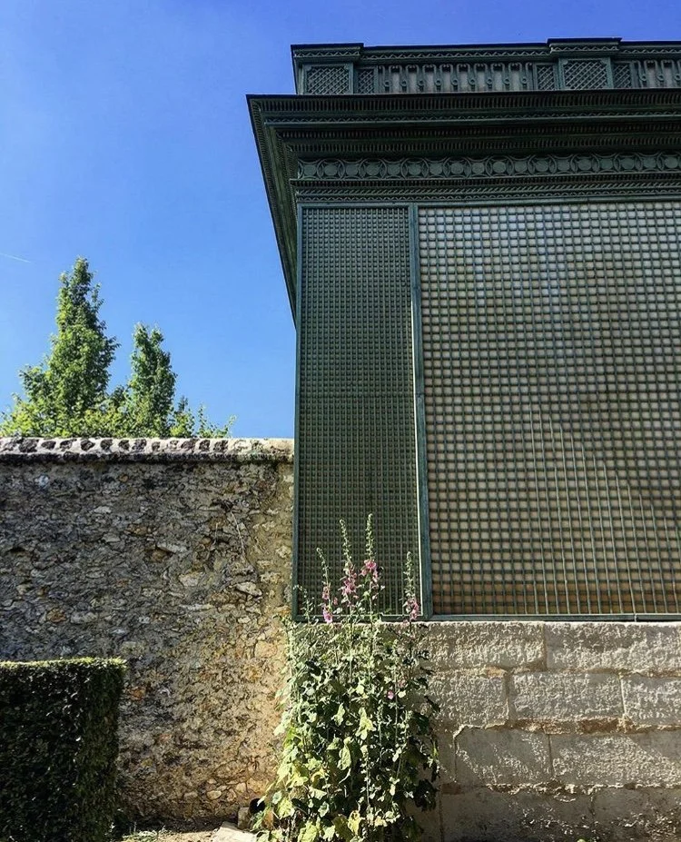 Close-up of the corner of a building with ornate metalwork, a stone wall, a bush with pink flowers, and a clear blue sky in the background.