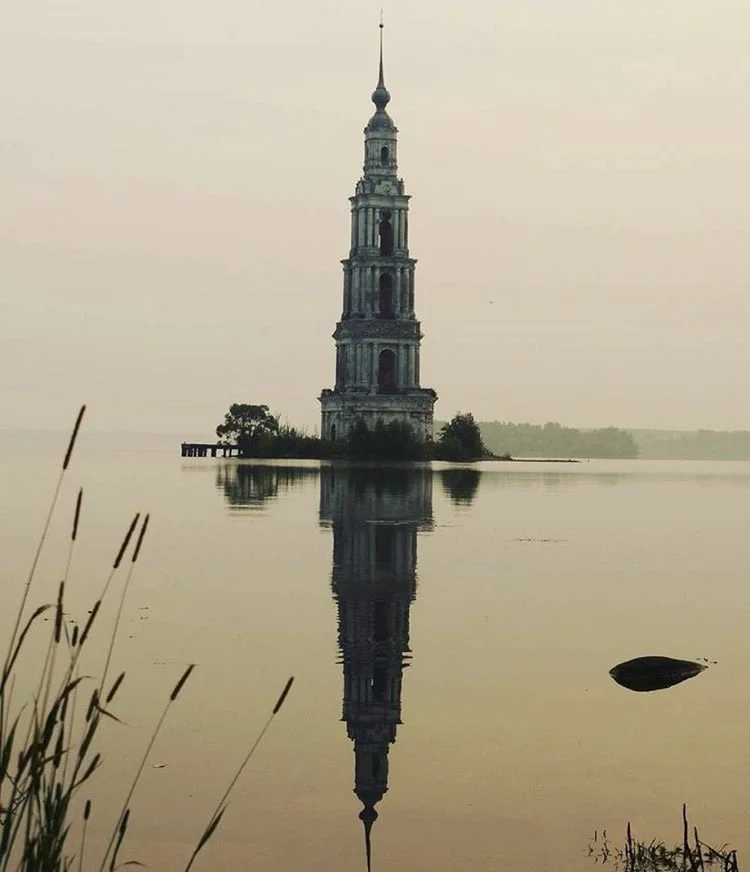A tall, historic tower or lighthouse reflected in calm water, with reeds and a rock in the foreground. The background is hazy with a distant shoreline.