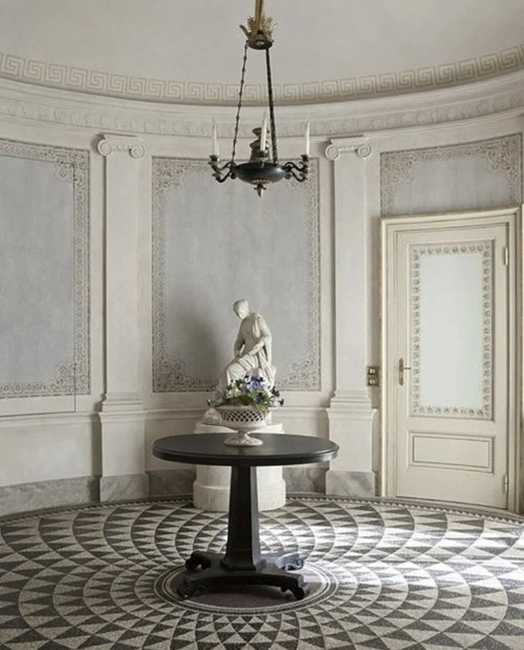 Elegant room with ornate white walls, a round dark wood table in the center holding a decorative flower arrangement, a classical white statue of a woman, and a black chandelier hanging from the ceiling. There is a white door on the right and intricat