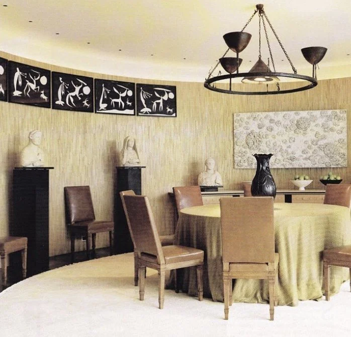 A round dining table with a beige tablecloth, surrounded by six brown chairs, in a room with beige walls. The room has white bust sculptures on pedestals along the wall, a large black vase on the table, and artwork on the wall. There is a black and g