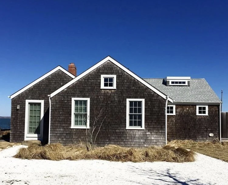 A house with gray shingle siding, multiple gabled roofs, and several windows, set against a clear blue sky.