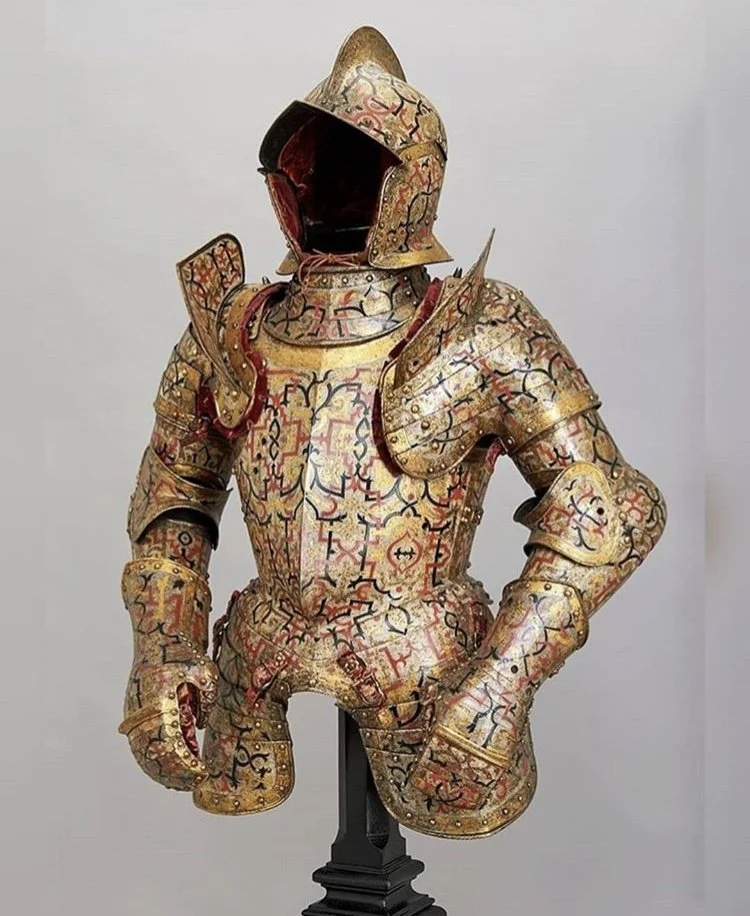 A suit of armor decorated with intricate red, black, and gold patterns with a matching helmet and shoulder guards, displayed on a black stand against a plain background.