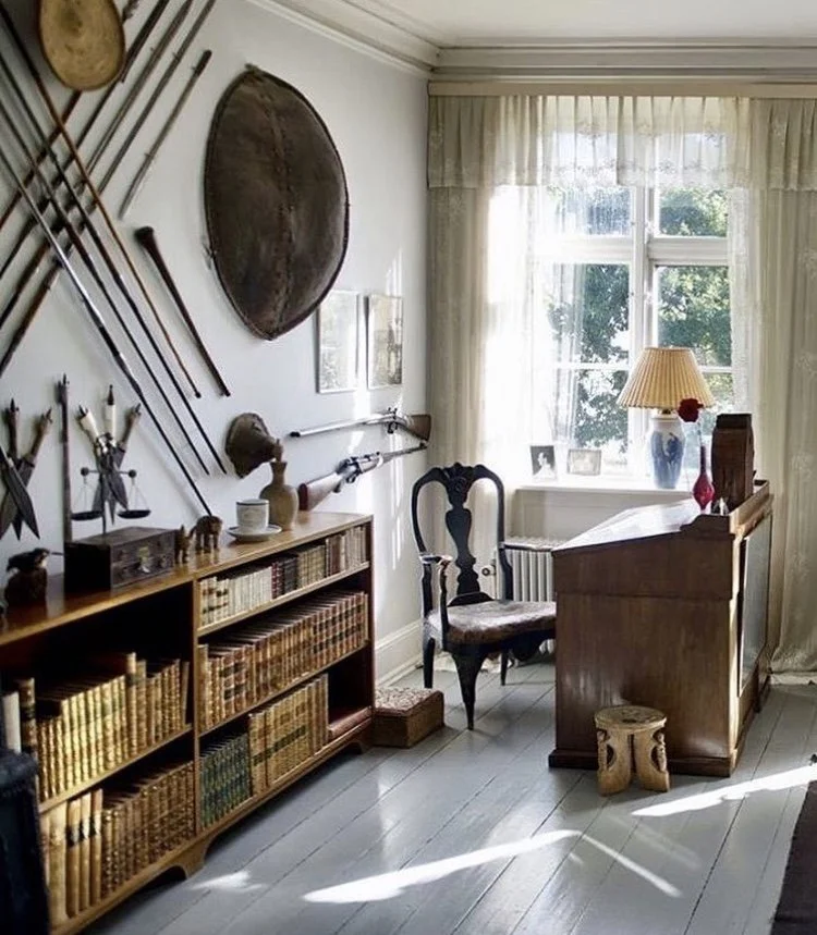 A cozy home office or study space with a wooden desk and a vintage chair by a window. The room features a bookshelf filled with old books, a collection of rifles and fishing poles mounted on the wall, and various decorative items including framed pic