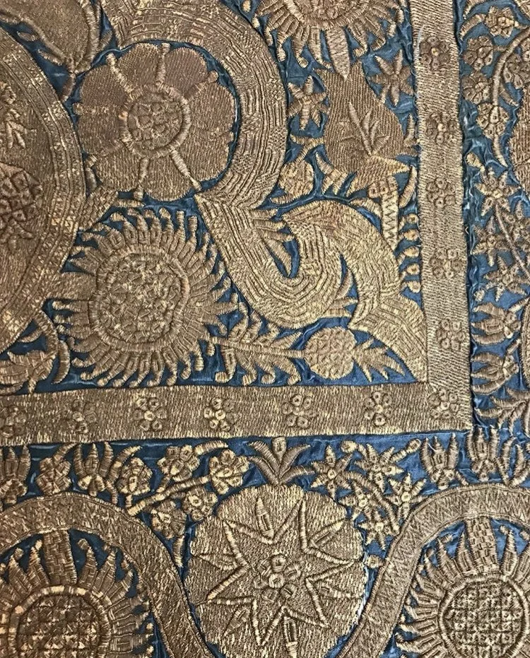 Close-up of an ornate, embroidered fabric with gold thread featuring floral and geometric patterns on a blue background.