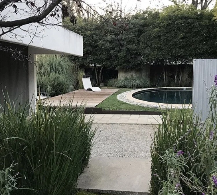 A backyard with a small pool, a wooden deck with a lounge chair, surrounded by lush green plants and trees.