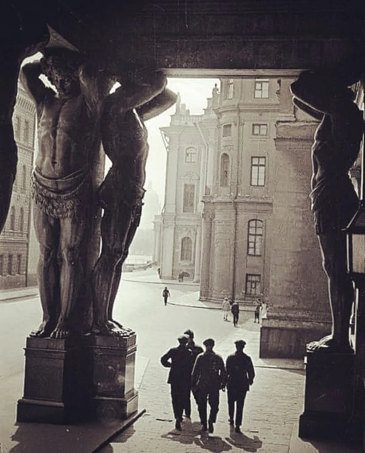 People walking under decorative statues of muscular male figures in an urban setting with historic buildings in the background.
