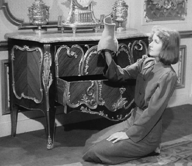 A woman is kneeling in front of an open piano, holding a vase above it, in a room with ornate furniture and decorations.
