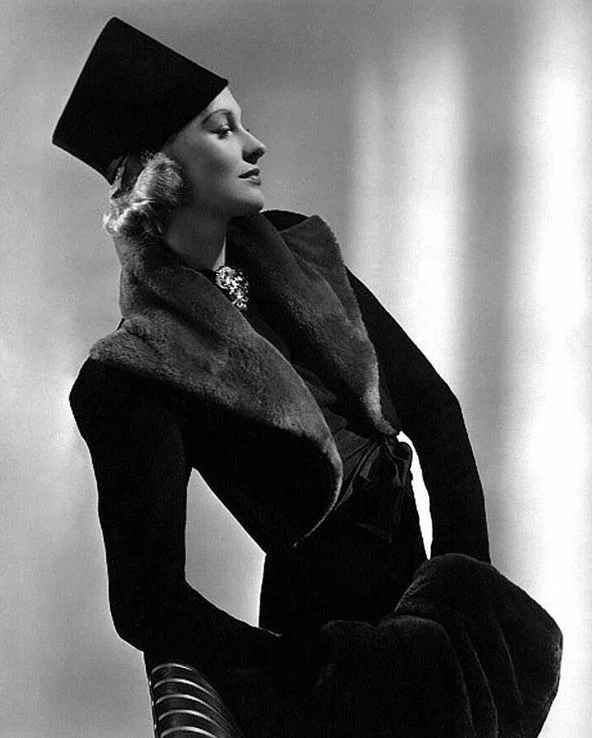 A black-and-white photo of a stylish woman in vintage fashion, wearing a fur shawl, an elegant hat, and jewelry, posing with a confident expression.