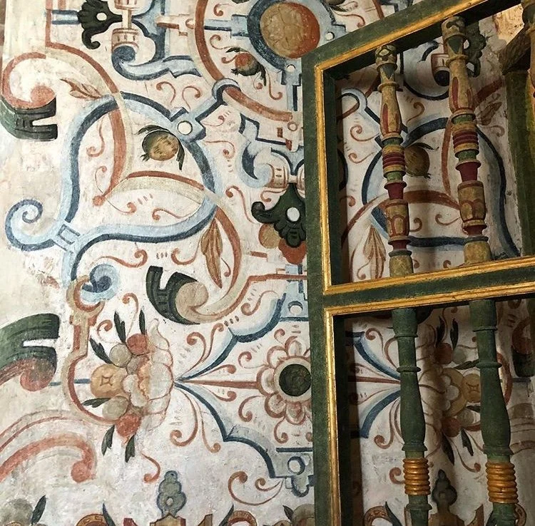 Close-up photo of a wall with intricate, colorful floral and geometric patterns, partially covered by a gold-framed mirror with decorative wooden spindles.
