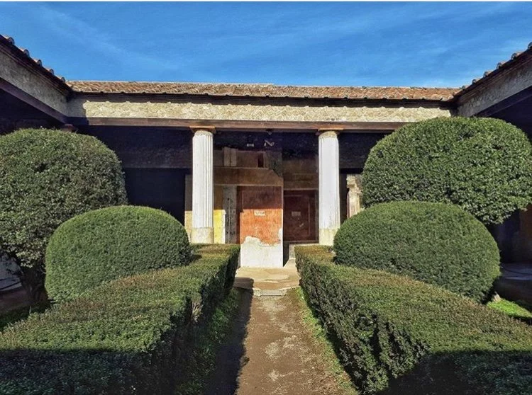 Front view of a building with four white columns, surrounded by lush, trimmed bushes and a pathway leading to the entrance.