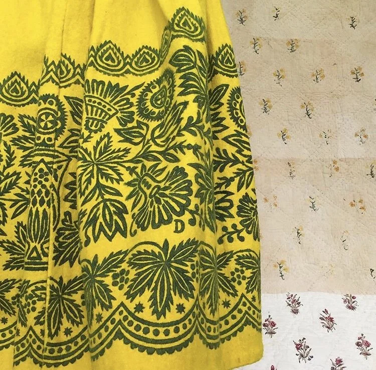 Yellow fabric with intricate dark green floral and geometric patterns, hanging against a background of wallpaper with small floral prints.