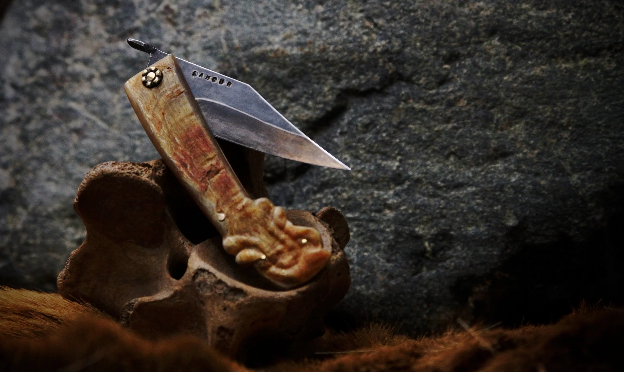 A folding knife with a handle made of wood and metal, resting on a skull-shaped piece of wood, against a stone background.